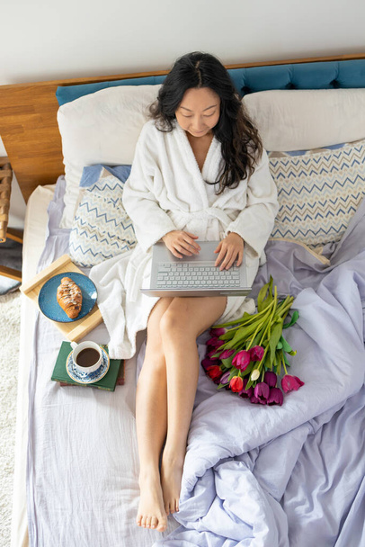 A young asian girl sits on the bed in the morning, uses a laptop, eats croissants and drinks coffee for breakfast. Enlarging a picture from the camera from top to bottom - Foto, afbeelding