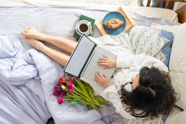 Μια νεαρή Ασιάτισσα κάθεται στο κρεβάτι το πρωί, χρησιμοποιεί ένα λάπτοπ, τρώει κρουασάν και πίνει καφέ για πρωινό. Μεγέθυνση μιας εικόνας από την κάμερα από πάνω προς τα κάτω - Φωτογραφία, εικόνα