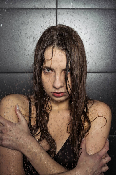 A beautiful brunette in a black swimsuit stands under pouring water droplets and poses against a black matte tiles. Wet skin and hair. - Фото, изображение