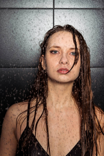 A beautiful brunette in a black swimsuit stands under pouring water droplets and poses against a black matte tiles. Wet skin and hair. - Fotó, kép