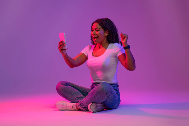 Online Win. Возбужденная черная женщина празднует успех со смартфоном в руке, радостная афро-американская женщина трясет сжатым кулаком и кричит да во время позирования за фиолетовый фон, скопировать пространство - Фото, изображение