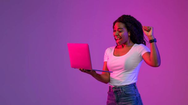 Radosna czarna kobieta trzymająca laptopa i świętująca sukces w świetle neonu, szczęśliwa młoda Afroamerykanka używająca komputera i podnosząca pięść z podnieceniem, stojąca nad fioletowym tłem - Zdjęcie, obraz
