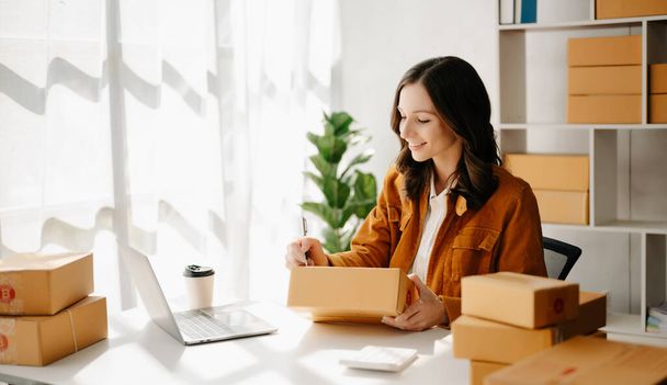 Startup small business SME, Entrepreneur owner woman using laptop taking receive and checking online purchase shopping order to prepare pack product boxes.  at home office - Foto, Bild