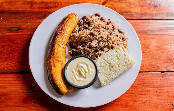 Gallopinto talíř se sýrem a maduro na dřevěném stole. Nikaragujský koncept jídla, tradiční gallo pinto jídlo s maduro a sýrem podávané - Fotografie, Obrázek