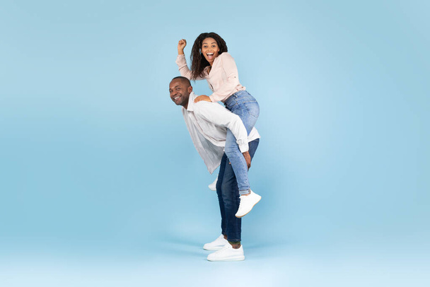 Overjoyed black man giving piggyback ride to his young wife, posing over blue studio background, carefree lovers enjoying time together, copy space, full length shot - Fotoğraf, Görsel