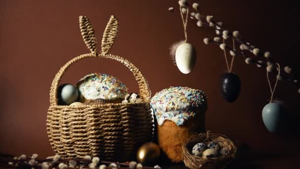 Easter holidays, cute basket with bunny ears on brown background. Easter cakes with colored sprinkles, colored golden eggs, and pussy willow decorations. Nest with eggs, Traditions - Felvétel, videó