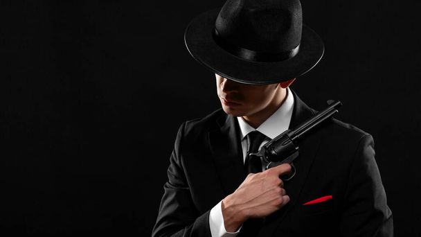 Gangster aus den 1940er Jahren mit Pistole. Mann in schwarzem Anzug und Hut mit Waffe vor dunklem Hintergrund. Foto mit Kopierraum. - Foto, Bild