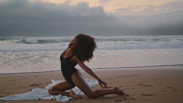 Sexy chica afroamericana bailando en la playa de arena nublado noche de verano. Morena rizada flexible haciendo elementos acrobáticos en un rendimiento apasionado. Mujer delgada en traje de baño negro cuerpo en movimiento con gracia - Foto, imagen
