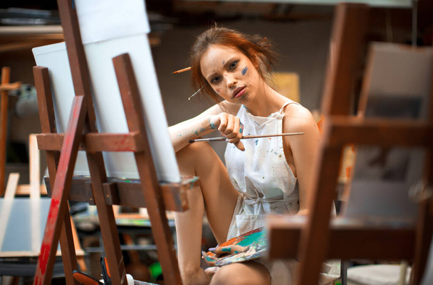 Портрет молодой художницы в студии. Она смотрит в камеру, думая о своих картинах. - Фото, изображение