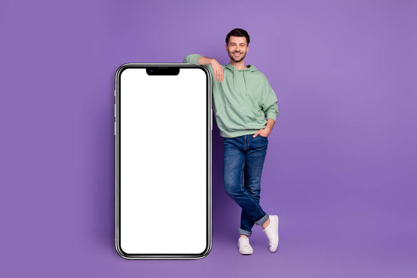 Full body photo of attractive young man leaning vertical white banner gadget dressed stylish khaki look isolated on purple color background. - Foto, Imagem
