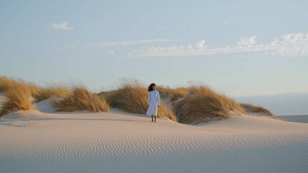 Jeune femme solitaire debout désert de sable devant haute herbe sèche le jour d'été. Fille afro-américaine bouclée portant une robe blanche posant en pleine nature sauvage à distance. Brune calme profiter du vent soufflant les cheveux. - Photo, image