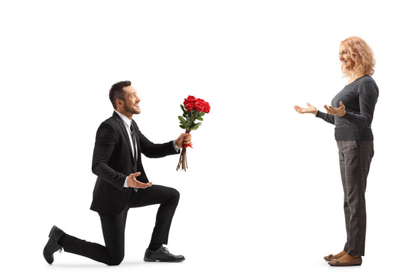 Fiatal férfi letérdel és egy csomó vörös rózsát ad egy nőnek, aki fehér háttérrel van elszigetelve. - Fotó, kép