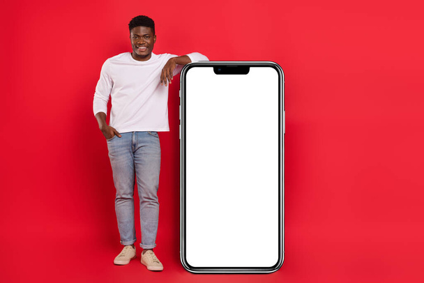 Full length photo of positive young male lean on big smartphone promotion board isolated on red color background. - Foto, imagen