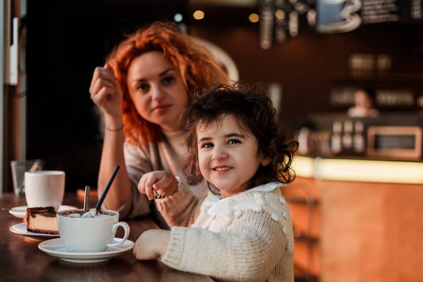 Schöne junge rothaarige Mutter mit niedlichen lockigen Tochter sitzen in gemütlichen Café und trinken heiße Schule. Muttertag. - Foto, Bild
