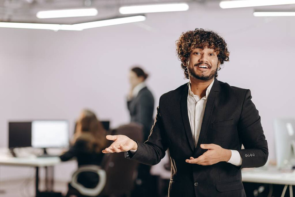 Excited businessman showing something. Smiling entrepreneur in black suit gesturing in office. Successful manager on blurred background. High quality photo - Photo, Image