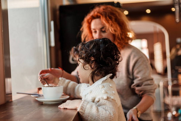 Όμορφη νεαρή κοκκινομάλλα μητέρα με χαριτωμένο σγουρά μαλλιά κόρη κάθονται σε ζεστό καφέ και πίνουν ζεστό σχολείο. Ημέρα μητέρων. - Φωτογραφία, εικόνα