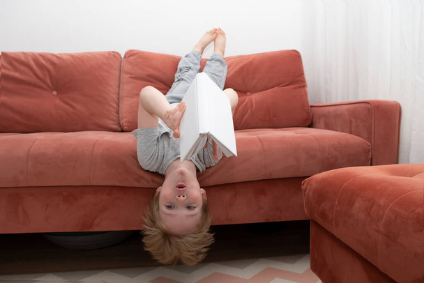 The boy is lying upside down on the sofa and reading a book in a white cover. How to teach a child to read and love books. Educational and entertaining childrens concept - Fotografie, Obrázek