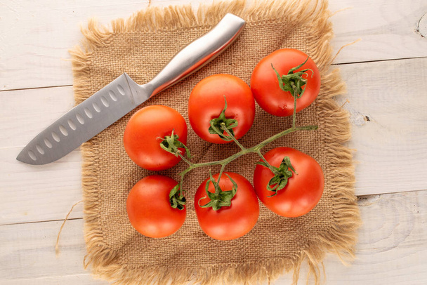 Several fresh tomatoes on branch with metal knife and jute napkin on wooden table, macro, top view. - Zdjęcie, obraz