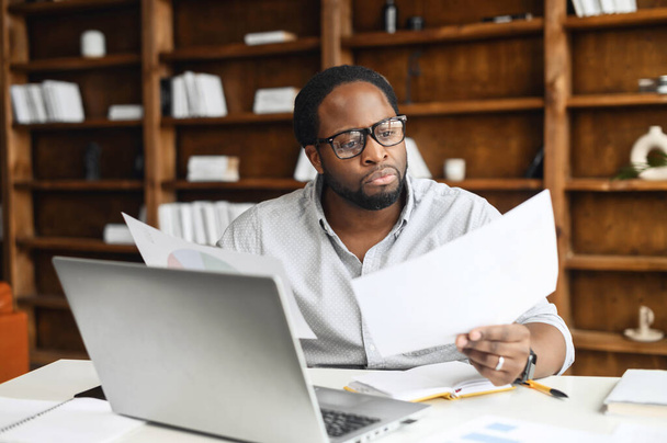 Clever and concentrated African-American guy is doing paperwork, a man looking through workpaper sitting at the desk with a laptop in office, a man checking report, collate data - Foto, Bild