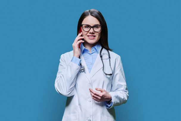 Young female doctor talking on mobile phone, on blue background of studio. Friendly physician talking to patient. Medical staff occupation, technologies mobile applications, health care medicine - Φωτογραφία, εικόνα