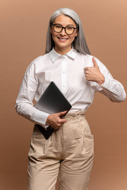 Waist up portrait of smiling asian mature woman holding closed laptop and showing big finger, while looking at camera with positive expression. Indoor studio shot isolated on beige background - Photo, image