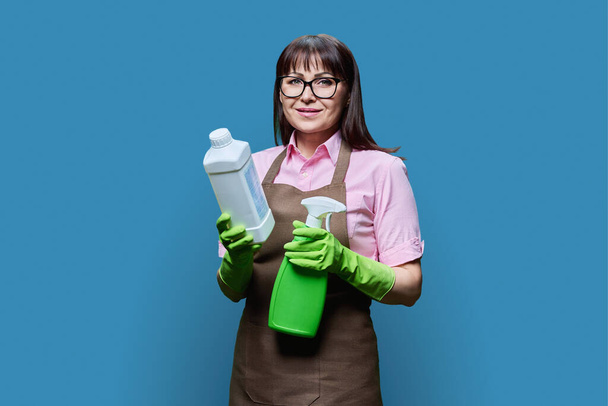 Middle aged woman in gloves apron with organic detergents, in hands looking at camera on blue color background. Home cleaning service, housekeeping cleanliness, natural eco organic detergents, ecology - Photo, Image