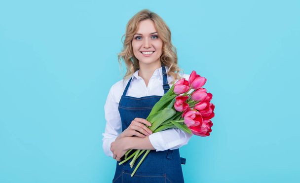 positive flower seller woman in apron with spring tulip flowers on blue background. - Фото, зображення