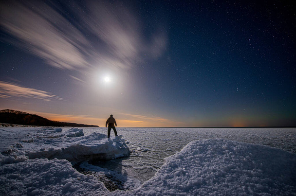 Traveler on the seashore covered with ice under a starry sky with clouds and moon - Foto, imagen