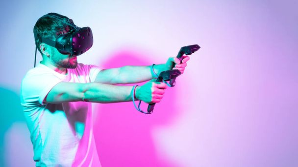 Bearded man in VR glasses plays in simulation figth and using controllers in in multicolored neon lights colorful background. Concept interactive online game in a virtual world. Copy space, banner. - Photo, Image