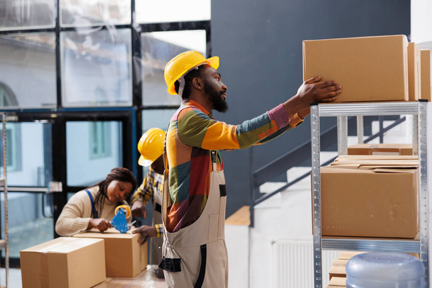 Warehouse package handler choosing cardboard box on shelf while standing on ladder. African american man storehouse worker preparing customer parcel and taking carton container - Photo, Image