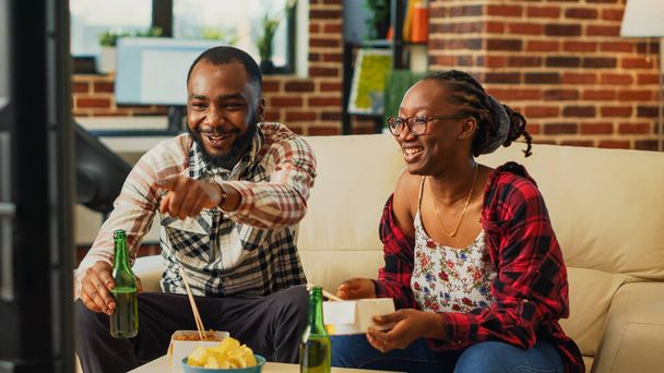 Afrikai amerikai pár ázsiai kaját eszik otthon, együtt töltik az időt elvitelre való étellel és sörösüvegekkel. Fiatal élettársak evőpálcikával tésztát esznek a nappaliban.. - Fotó, kép