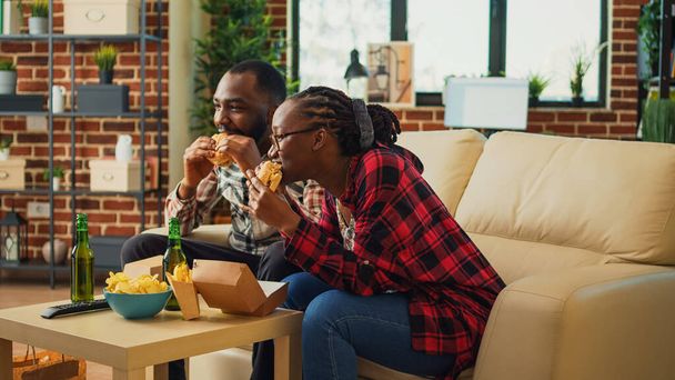 Cheerful young people eating hamburgers and fries in living room, watching favorite film on tv and drinking alcohol. Boyfriend and girlfriend serving fast food order from takeout place. - Foto, Bild
