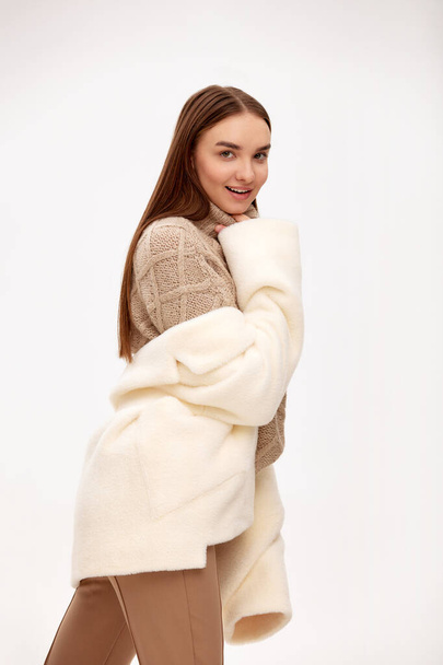 Porträt eines schönen Mädchens im modischen Schaffell-Mantel und Strickmütze, das auf dem Hintergrund von Wintertannen steht. Wintermode. Kopierraum - Foto, Bild