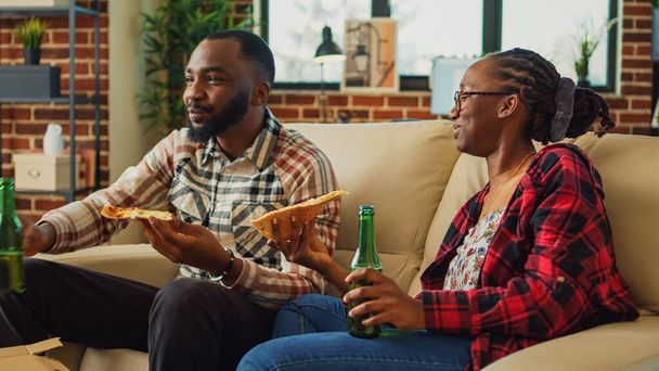 テレビの前でピザのスライスを食べるアフリカ系アメリカ人、自宅でお気に入りのショーを見て大きな。現代のリラックスしたカップルは、ファーストフードやビールのボトルを提供しています. - 写真・画像