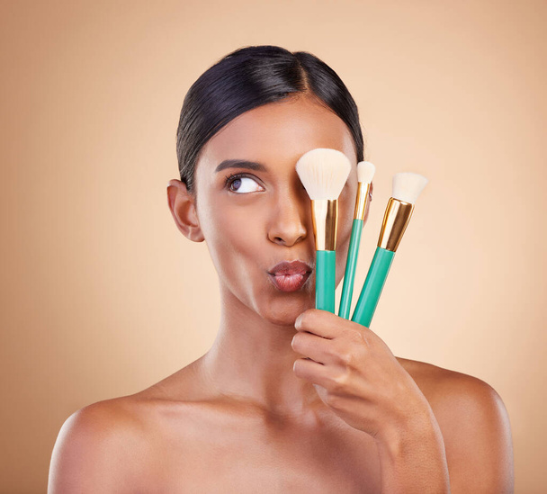 Skincare, brush and Indian woman with makeup, foundation and funny on brown studio background. Female, lady or cosmetics tools with glamour, elegant or beauty for luxury grooming or facial expression. - Photo, Image