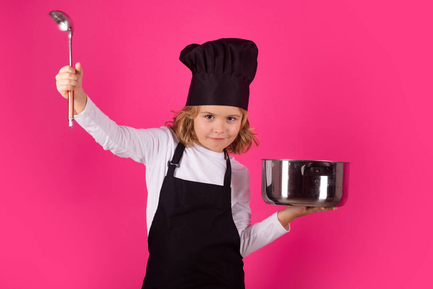 Детский повар готовит с кастрюлей и ковшами. Ребенок в форме плиты и шляпе шеф-повара готовит еду на кухне, студийный портрет. Концепция приготовления пищи, кулинарного и детского питания - Фото, изображение