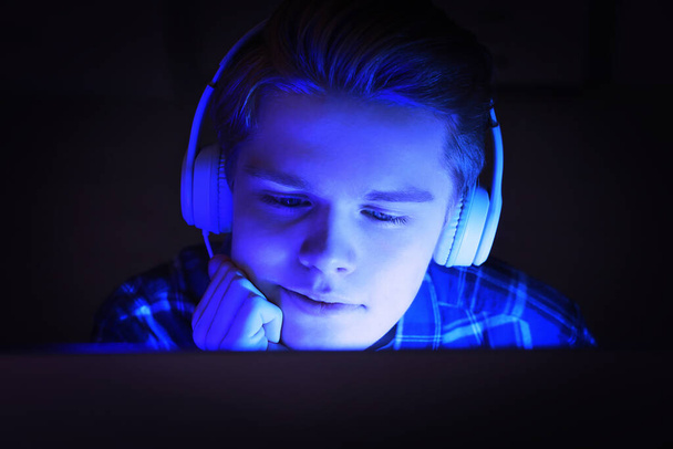 Internet addiction. Teenage boy in headphones using device at night. Toned in blue - 写真・画像