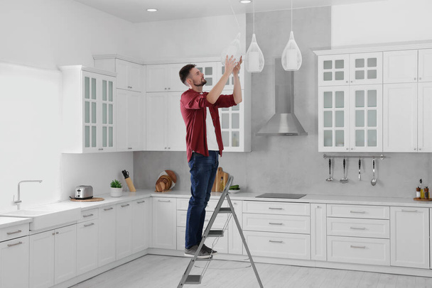 Young man installing ceiling lamp on stepladder in kitchen - Fotografie, Obrázek