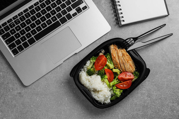 Container with tasty food, laptop, cutlery and notebook on light grey table, flat lay. Business lunch - Foto, afbeelding