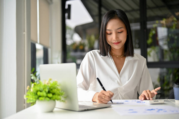 Profesyonel ve çekici Asyalı iş kadını finansal raporlarla ilgili bir şeyler yazıyor, satış verilerini analiz ediyor, ofisindeki masasında çalışıyor.. - Fotoğraf, Görsel