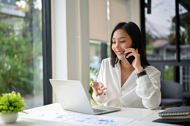 Çekici ve profesyonel Asyalı iş kadını iş ortağıyla telefonda konuşuyor, telefonda iş anlaşması yapıyor, ofisinde çalışıyor.. - Fotoğraf, Görsel