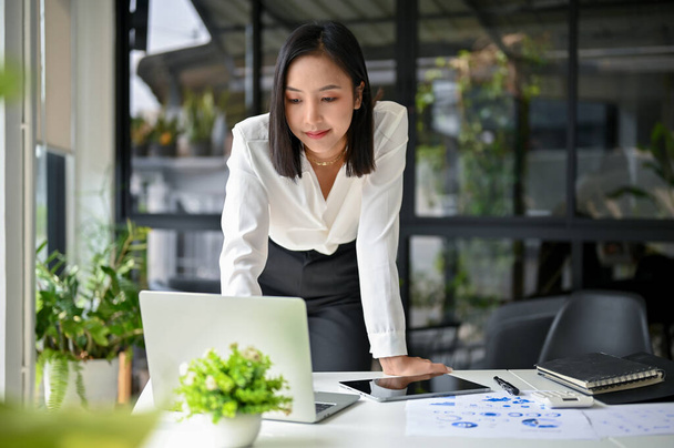Profesjonalne i wspaniałe tysiąclecia Azjatyckie businesswoman pracuje nad swoim biznesowym projektem inwestycji finansowych, opierając się na stole i za pomocą laptopa. - Zdjęcie, obraz