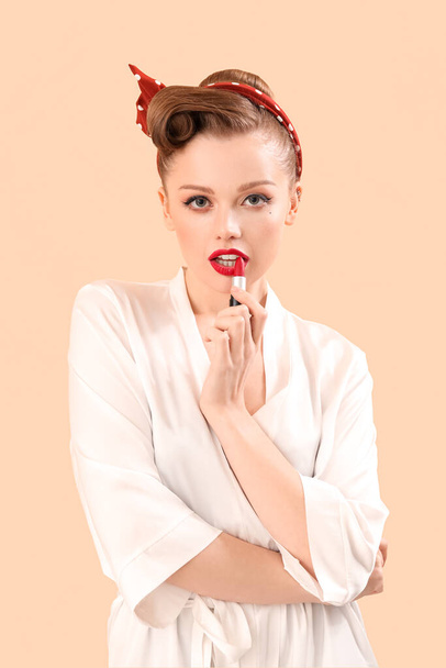 Young pin-up woman applying red lipstick on beige background - Photo, Image
