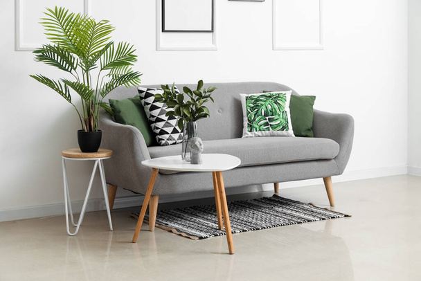 Gemütliches Sofa mit Kissen und Zimmerpflanzen auf Tischen neben weißer Wand - Foto, Bild