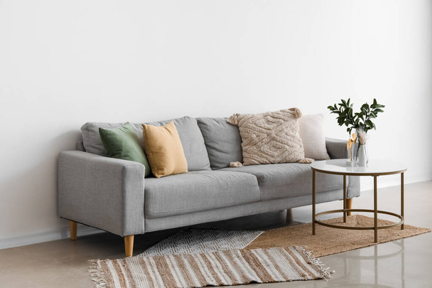 Canapé confortable gris avec coussins et branches de plantes dans un vase sur la table près du mur clair - Photo, image