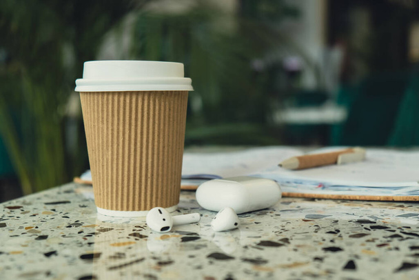 Take away coffee in craft recycling paper cup with paper notebook with wireless headphones. Mockup Coffee break. Audio healing, sound therapy wellness rituals, positive mental health habits listening - Photo, Image