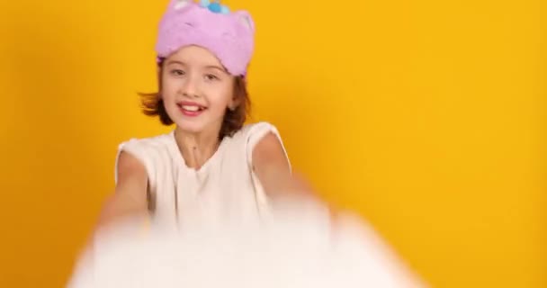 Happy teenage girl in white pyjamas with a violet sleeping mask hold pillow and fight in the studio on a yellow background - Footage, Video