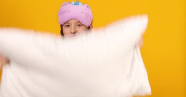 Funny teenage girl in white pyjamas with a violet sleeping mask hold pillow, having fun, make faces in the studio on a yellow background - Footage, Video