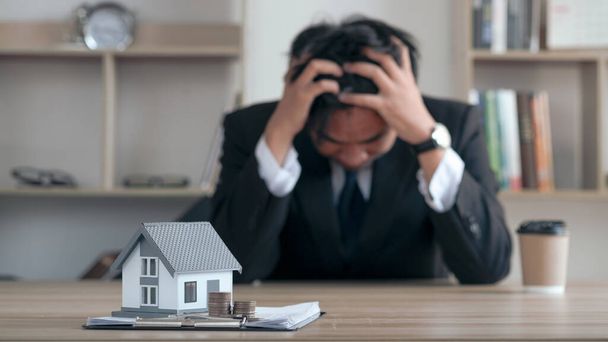 Concept of real estate debt spreading around the world.world economic crisis. recession, inflation. Businessman sitting stressed holding his head with the house model and contract document in front. - Фото, изображение