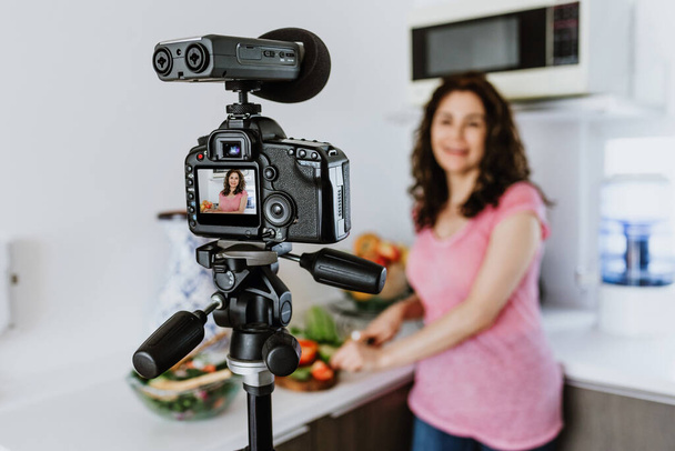 Latin woman influencer recording video class or live stream with camera about healthy eating and nutrition in kitchen at home in Mexico, hispanic people - Photo, Image